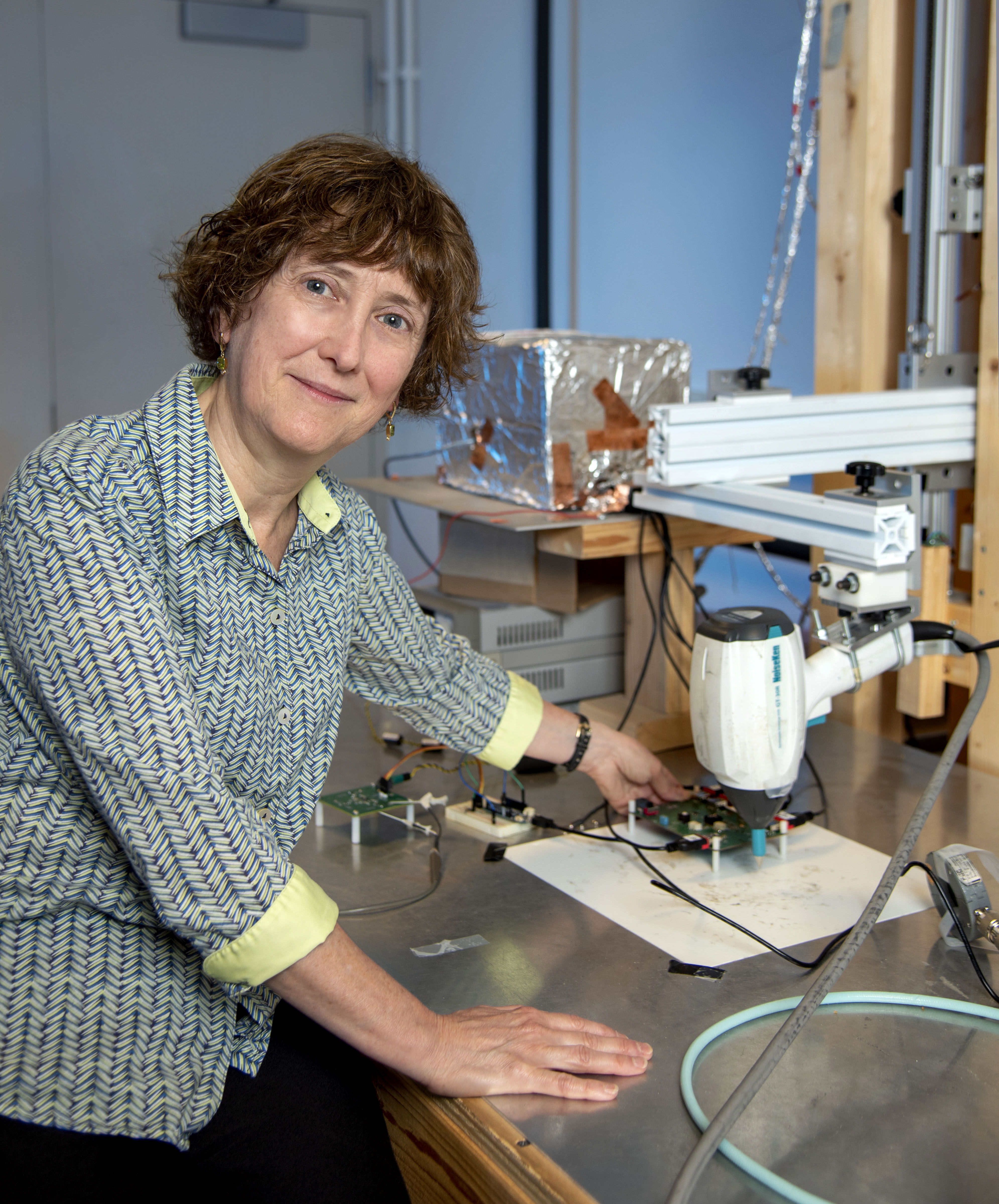 A woman stands at a laboratory counter with science equipment around her. She looks at the camera and smiles